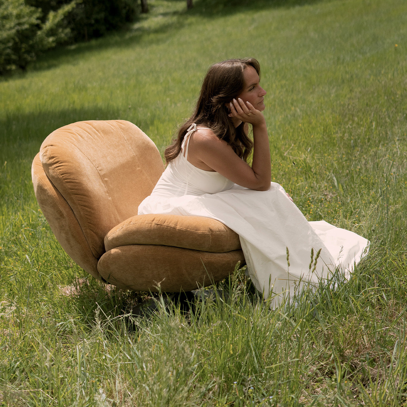 Fauteuil lounge arrondi en velours camel posé dans l'herbe, femme en robe blanche assise, ambiance naturelle – Maison Pavane