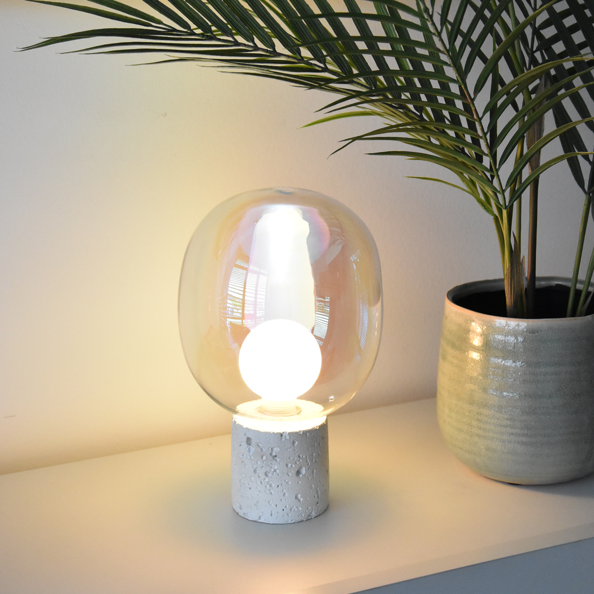 Lampe à poser Bulle en verre irisé et béton blanc, allumée sur une étagère blanche à côté d'un palmier dans un pot en céramique