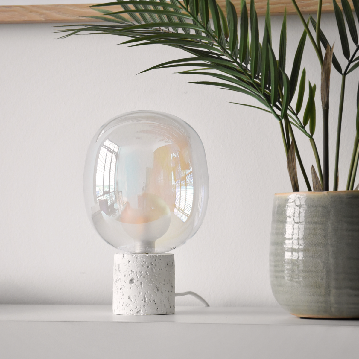 Lampe à poser Bulle en verre irisé et béton blanc, posée sur une étagère blanche près d'un palmier en pot devant un mur blanc