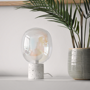 Lampe à poser Bulle en verre irisé et béton blanc, posée sur une étagère blanche près d'un palmier en pot devant un mur blanc