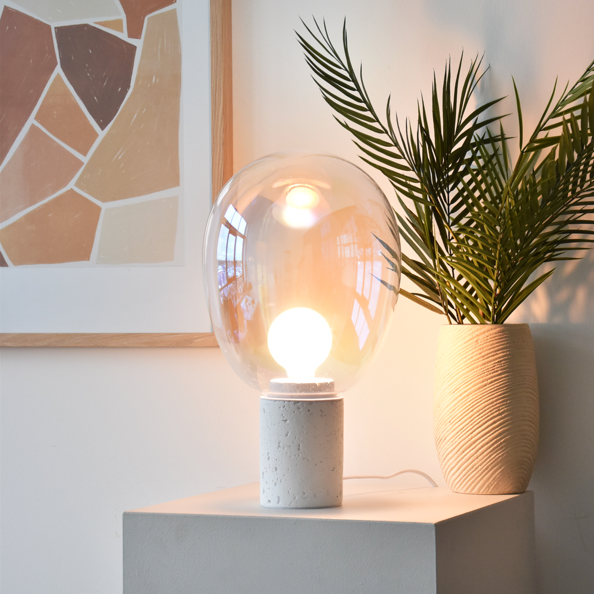 Lampe à poser Bulle en verre irisé et béton blanc, allumée sur un meuble blanc près d'un palmier en pot et d'un cadre aux tons terracotta