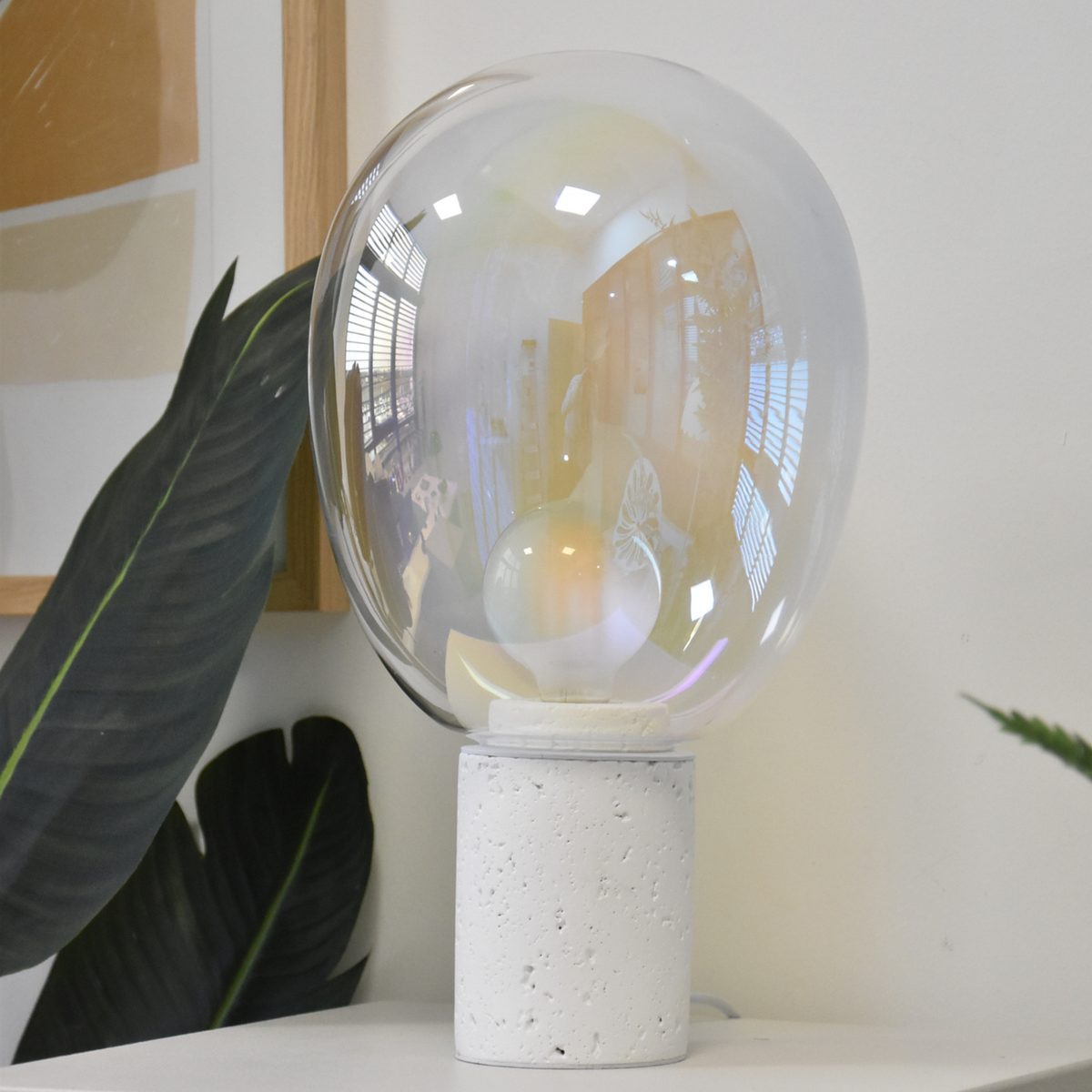 Lampe à poser Bulle en verre irisé et béton blanc, posée sur une étagère blanche devant un cadre en bois et une grande plante verte