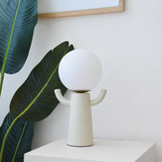 Lampe Paul en céramique et verre opalin blanc cassé éteinte, posée sur un socle blanc devant une plante tropicale et un cadre en bois