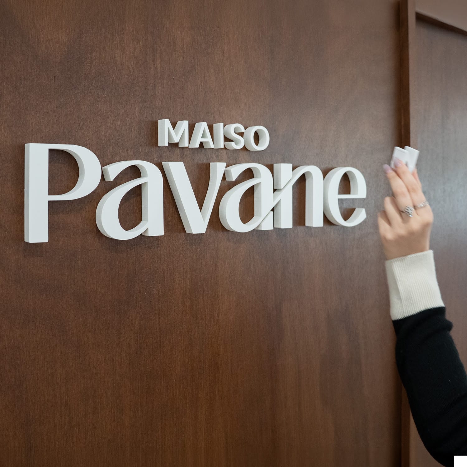 Logo Maison Pavane en lettres blanches relief sur panneau bois brun, main posant la dernière lettre – Maison Pavane
